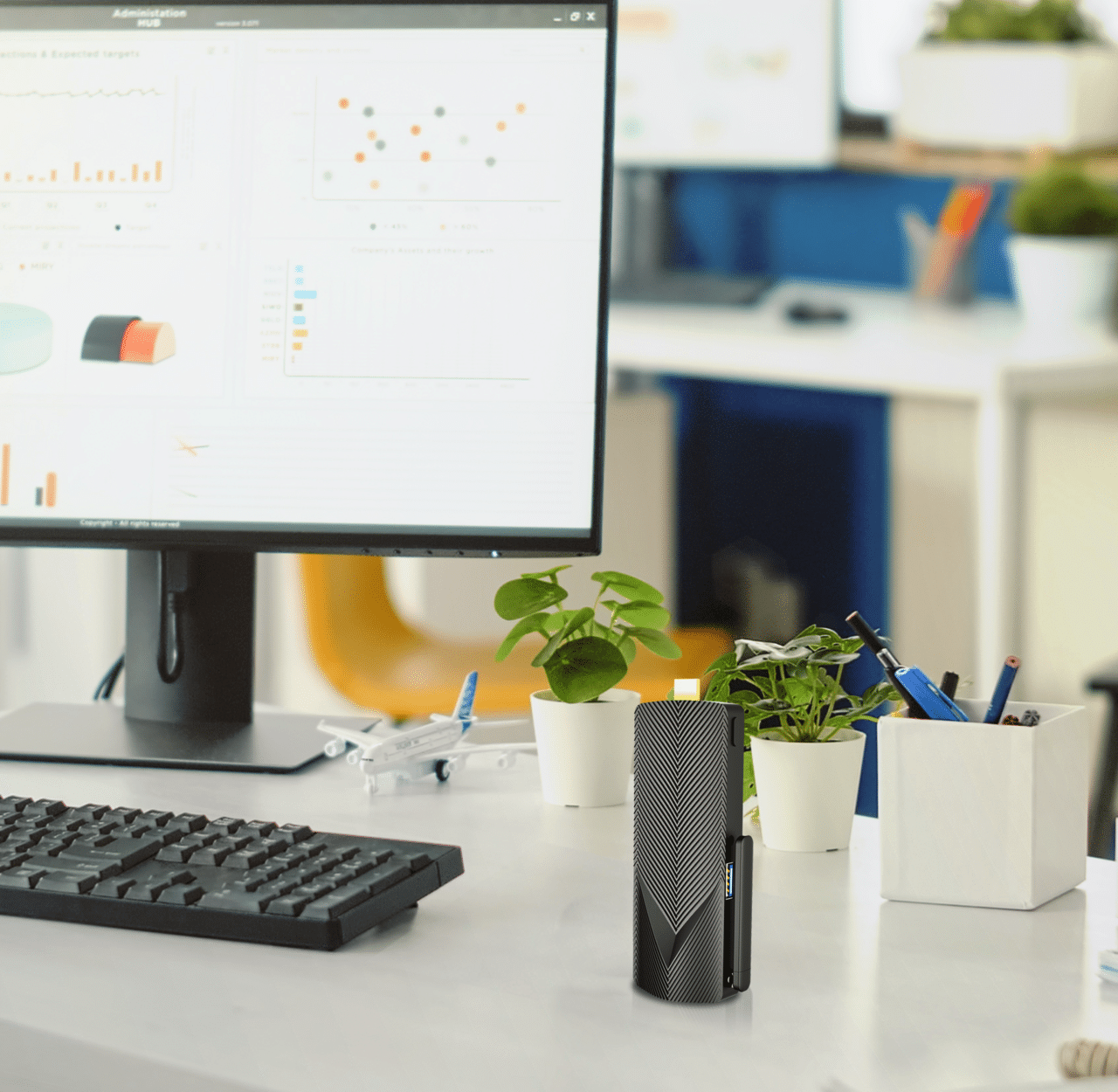 A workspace featuring a computer monitor displaying graphs, a mini a mechanical keyboard, office supplies, with potted plants adding a touch of greenery. This setup includes mini PCs for an efficient use of space